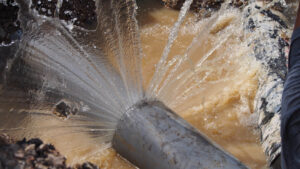 Close up of a burst pipe leaking water, sitting in muddy water.
